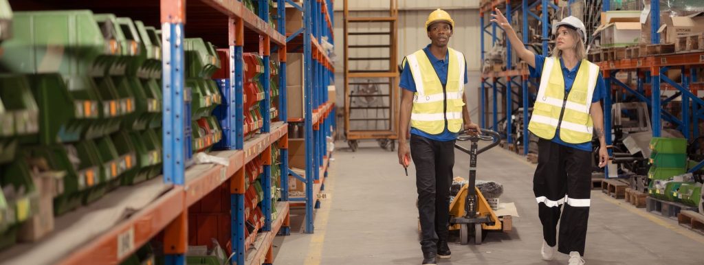 Two warehouse workers wearing reflective safety vests and hard hats walk through an aisle between tall metal shelving units stocked with boxes, with a hand pallet truck visible in the background.