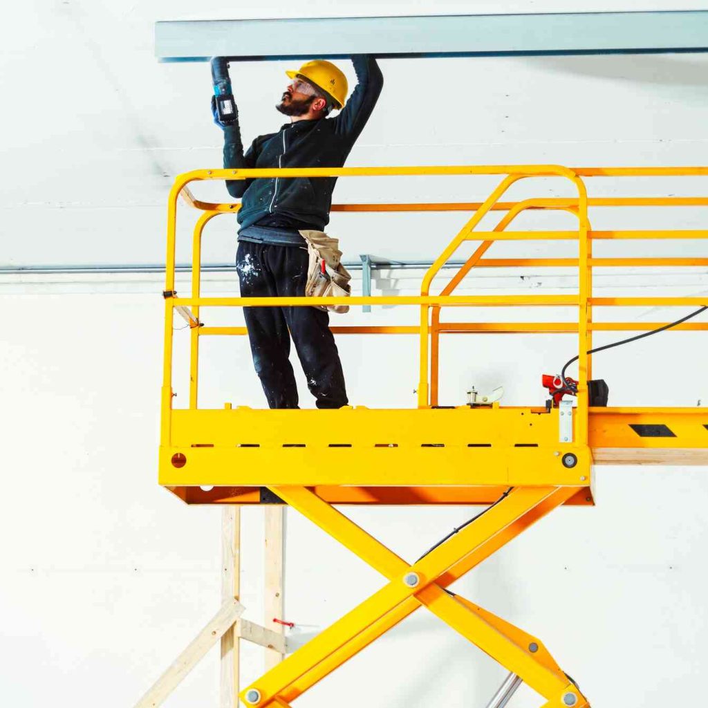 A construction worker in safety gear stands on a bright yellow aerial work platform, drilling into the ceiling with a power tool.