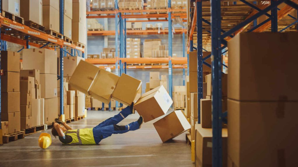 Shot of a Warehouse Worker Has Work Related Accident. He is Falling Down BeforeTrying to Pick Up Heavy Cardboard Box from the Shelf. Hard Injury at Work.