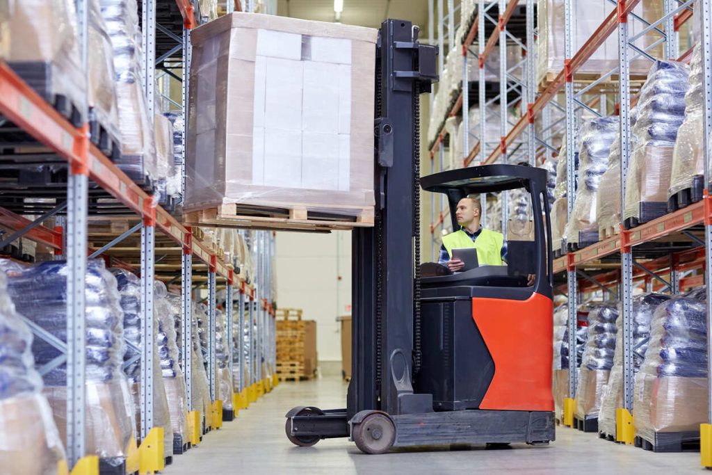 A warehouse worker wearing a safety vest operates an electric pallet stacker to move a loaded pallet down an aisle between tall metal shelving units stocked with wrapped and palletized goods.